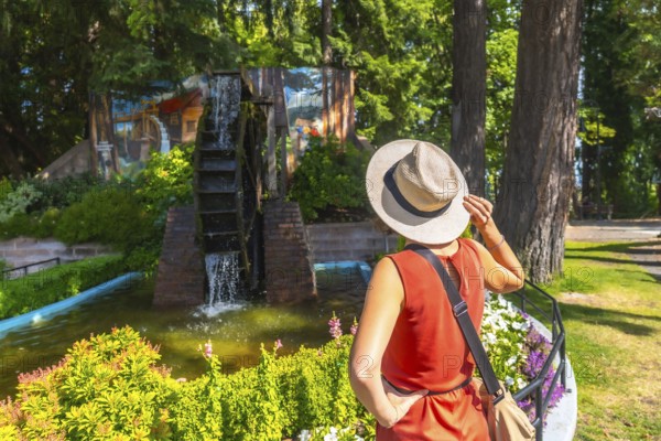 Tourist wearing a straw hat holds the brim while admiring a water wheel in a garden setting in