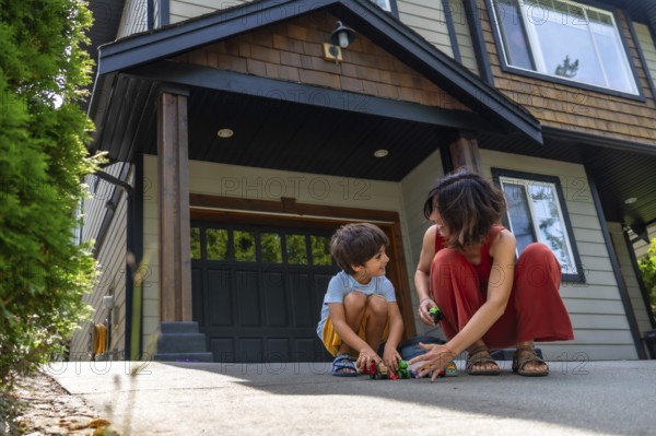 Mother and her young son are happily playing with toy cars on the driveway of their modern two