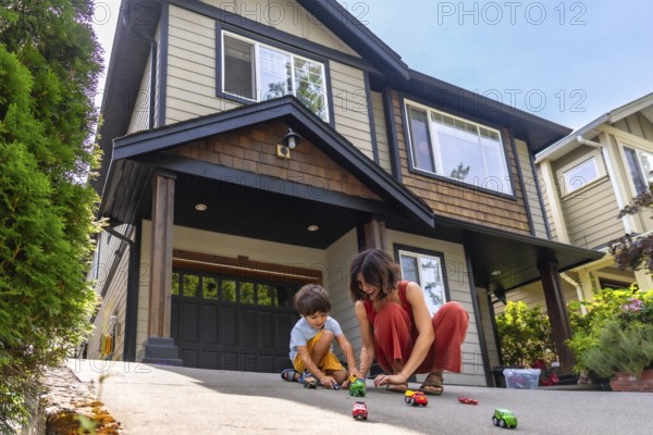 Mother and her young son are happily playing with toy cars on the driveway of their suburban home,