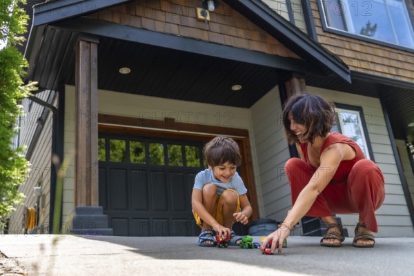 Mother and her young son are happily playing with toy cars on the driveway of their modern suburban