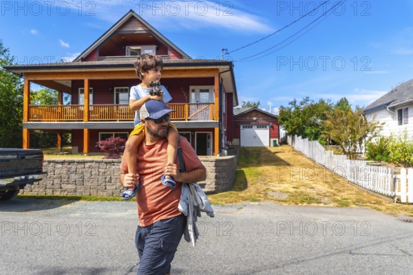Father carrying son on shoulders while walking past a two story house with a porch and detached