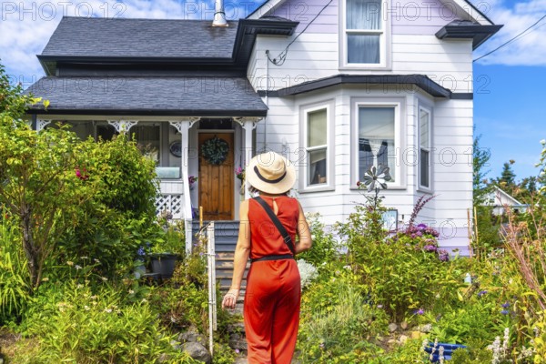 Tourist wearing a straw hat and an orange jumpsuit walks up a garden path towards a charming house
