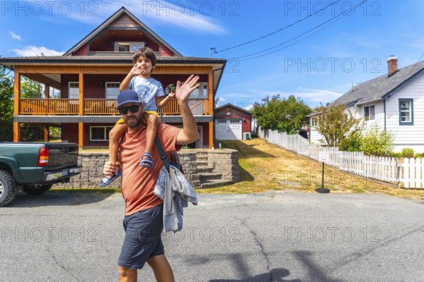 Father carrying his son on his shoulders waving in front of their house in chemainus, vancouver