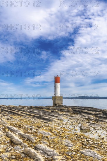 Red and white lighthouse standing on a rocky beach with a blue sky and clouds in the background in