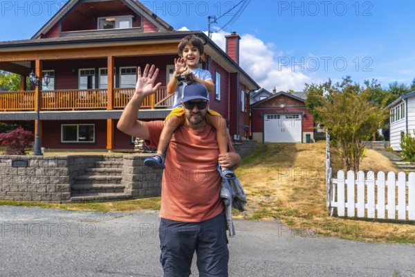 Father carrying his son on his shoulders, both waving, standing in front of their house in