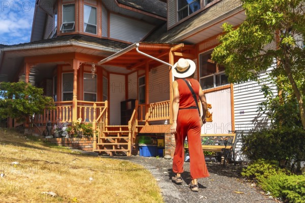 Tourist in a straw hat and red jumpsuit walking away from a colorful victorian house in chemainus,