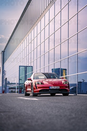 Red car standing in front of a modern glass building, reflecting sky in the background, Porsche
