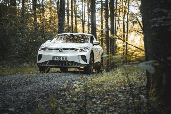 A car is parked in a forest on a path surrounded by autumn leaves and sunlight, VW ID4 electric