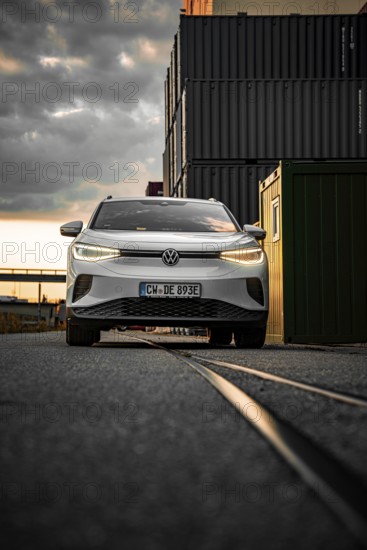 White car drives past container warehouse at sunset, dramatic urban scene, VW ID4 electric car,