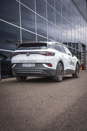 White car in front of glass façade on paved road, VW ID4 electric car, Deer Carsharing, Calw,
