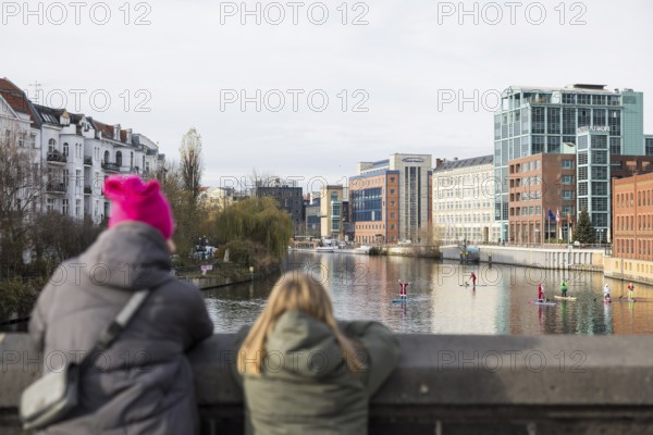 Passers-by watch water sports enthusiasts disguised as Santa Claus with SUPs on the Spree on the