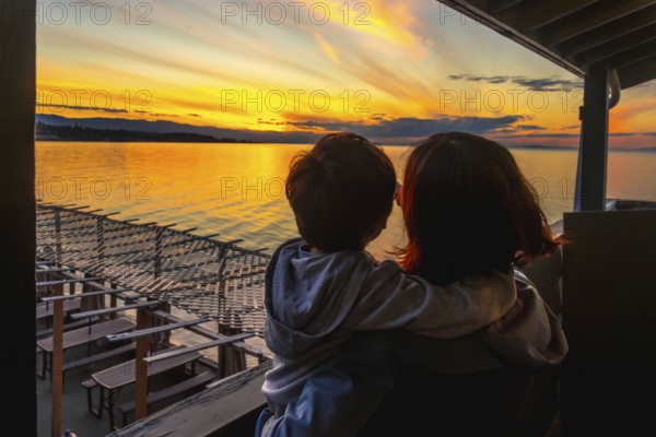 Mother and child share a tender moment, gazing at a vibrant sunset over the tranquil waters of