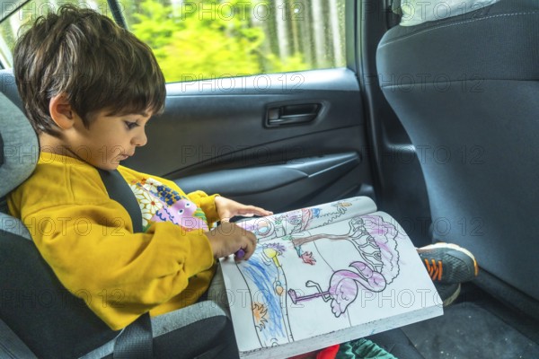 Young child concentrating on a coloring book in a car seat during a scenic drive through the