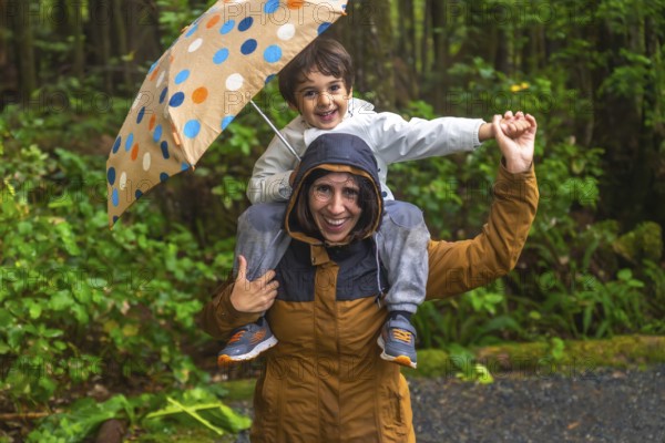 Mother carries her smiling son on her shoulders under a colorful umbrella, both wearing raincoats