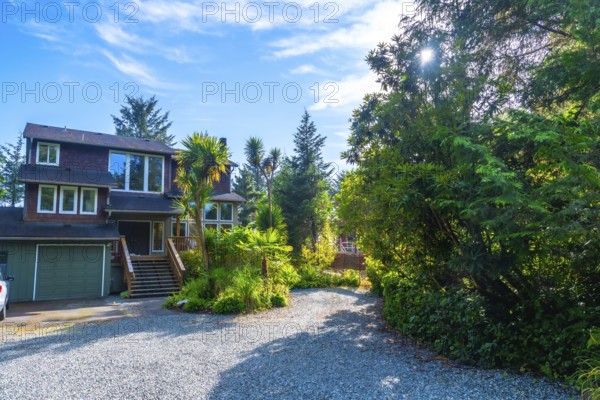 Lush green vegetation surrounds a charming two story house with a gravel driveway and attached