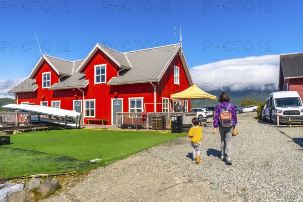 Mother and son are walking on a gravel path near a vibrant red house with white trim, canoes