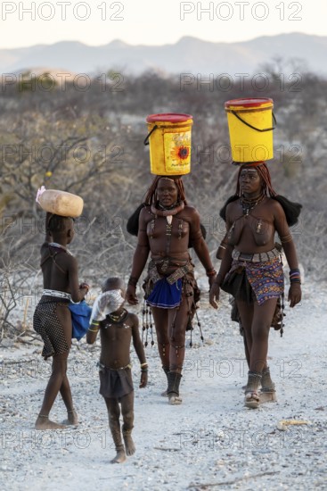 Himba woman and children run to fetch water with canisters through dry countryside, traditional