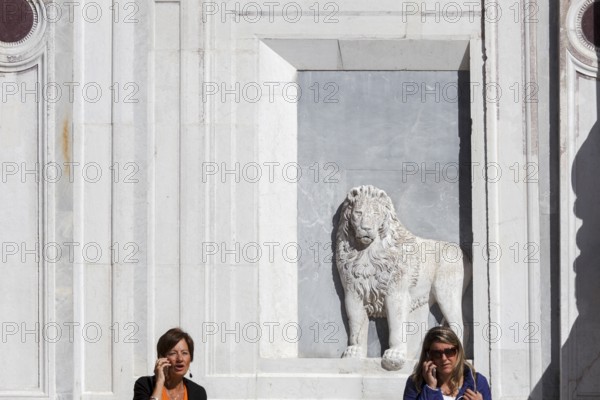 2 woman make phone calls in front of the lion relief on the façade of the Scuola Grande di San