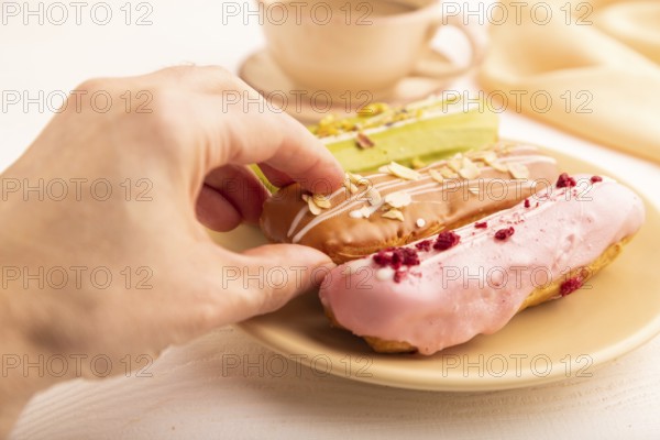Glazed Eclairs on white wooden background and orange linen textile with hand, cup of coffee, side