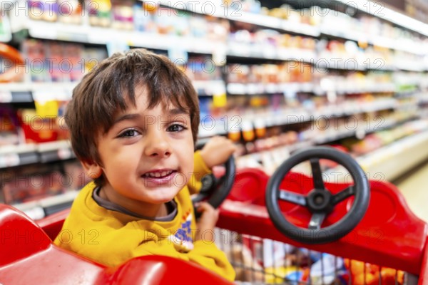 Cheerful child is enjoying a shopping trip, sitting in a shopping cart with a toy steering wheel