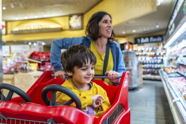 Mother pushing a shopping cart with her son sitting inside, enjoying their grocery shopping