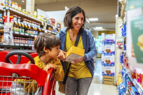 Smiling mother holding a yellow box while shopping with her son sitting in a shopping cart in a