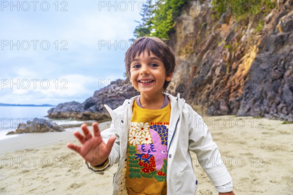 Happy child waving on a sandy beach with rocky cliffs and ocean in the background, enjoying a sunny