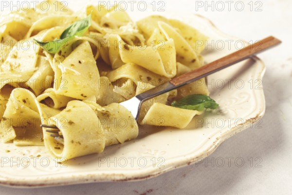 Fresh papardelle pasta, with basil and pesto sauce, is served, on a white plate, with a fork held