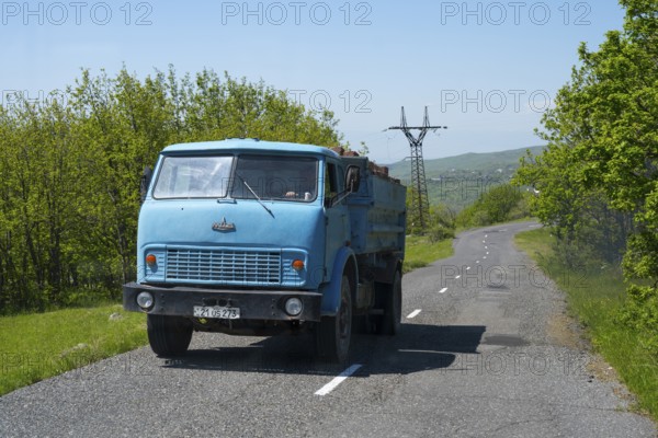 A blue truck drives on a sunny country road surrounded by trees and a power pole, MAZ truck,
