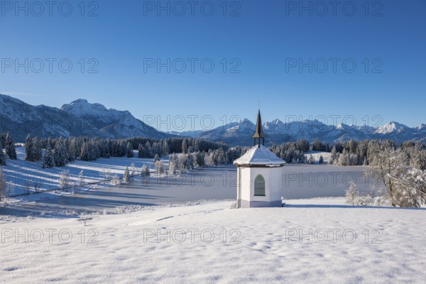 Small chapel in front of snow-covered mountains and forests under a clear sky, Hegratsrieder See,