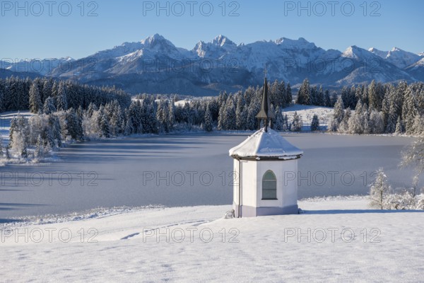 Chapel on the shore of a frozen lake surrounded by snow and mountains under a blue sky,