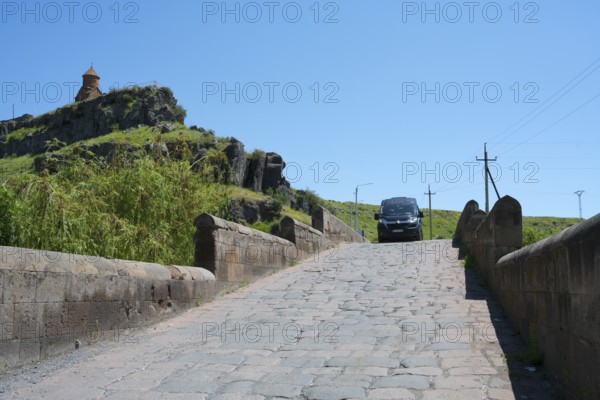 A car drives over a stone bridge with a building on a green hill in the background under clear sky,