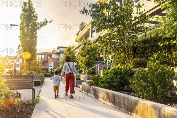 Mother and son holding hands are walking away from the camera on a sidewalk in a residential