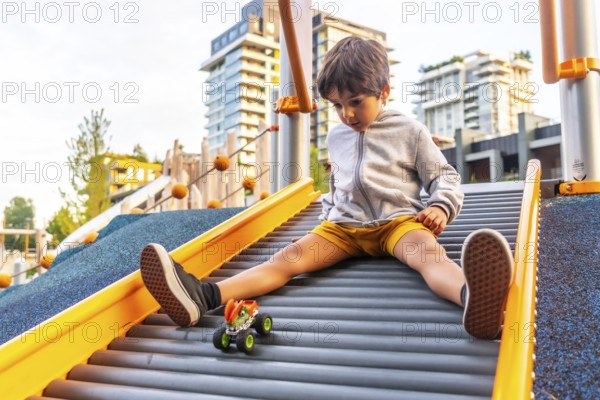 Young boy sitting on a playground slide, gazing at his toy monster truck while enjoying a sunny day