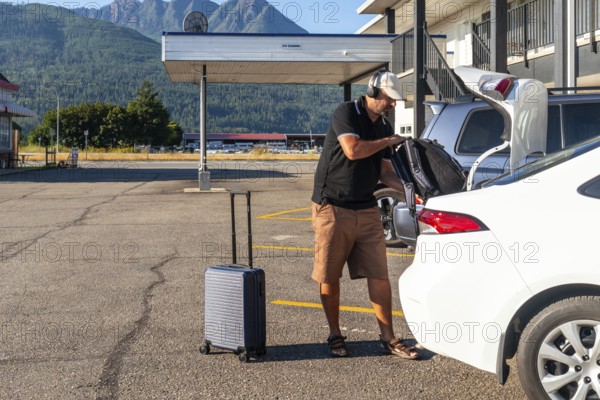 Tourist wearing headphones is loading luggage into car trunk in motel parking lot, preparing for