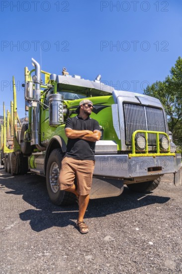 Truck driver confidently posing with arms crossed, leaning against a modern green logging truck in
