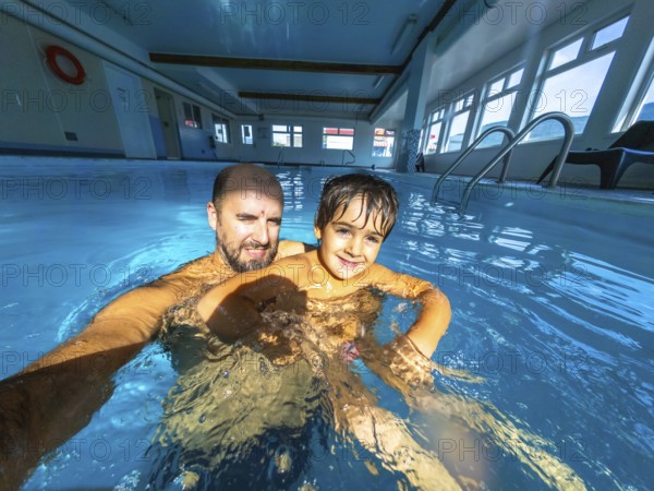 Father and son enjoying a playful moment while taking a selfie in an indoor swimming pool, creating