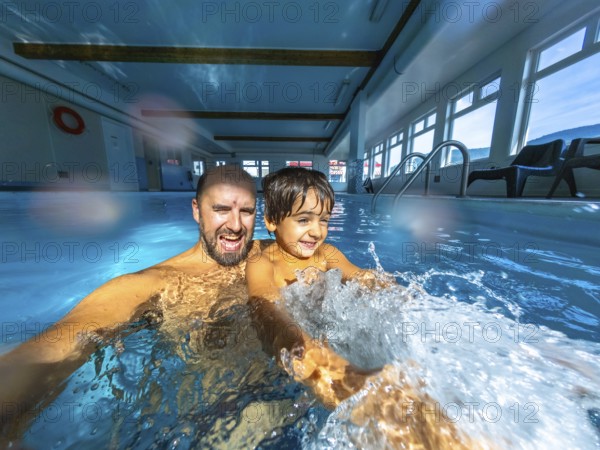 Father and son enjoying playful moments while splashing in an indoor swimming pool, creating a
