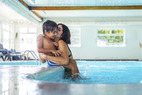 Mother holding and kissing her son at the poolside, with his feet splashing in the clear water,