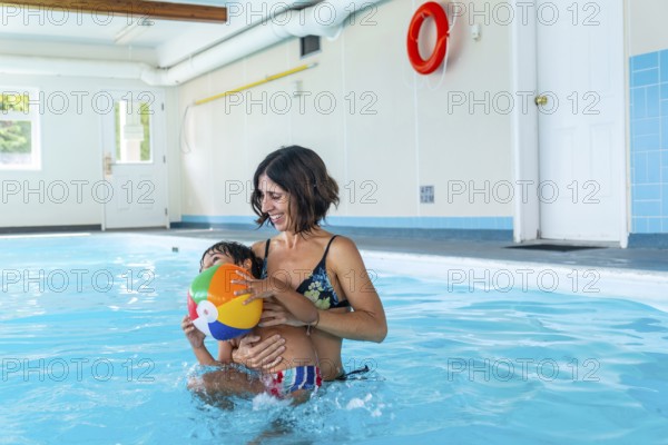 Mother holding her son while playing with a vibrant inflatable beach ball in an indoor swimming