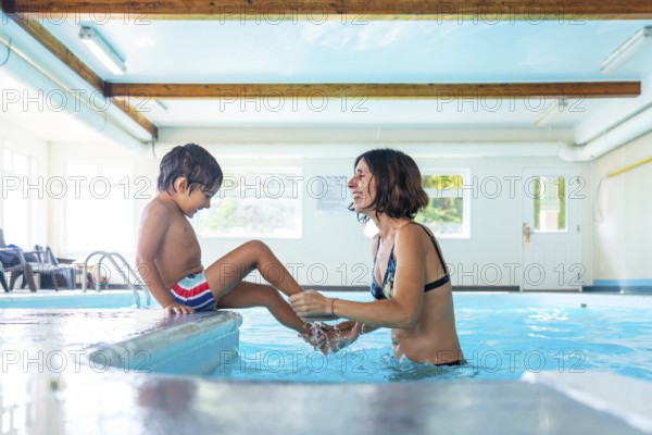 Happy mother and son enjoying playful moments at the edge of an indoor swimming pool, splashing