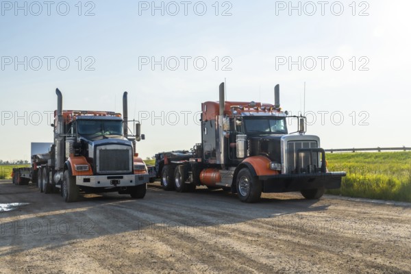 Two powerful semi trucks, gleaming in orange and black under a clear sky, parked on a dirt road