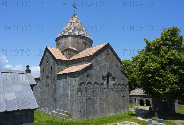 Medieval stone church with dome and cross under blue sky next to green trees, Sanahin monastery,