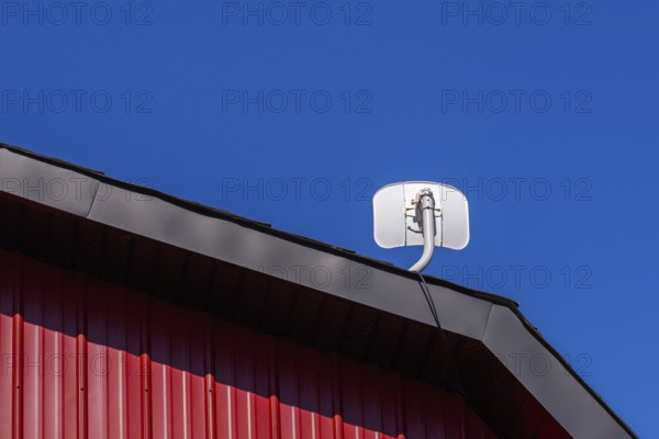 White satellite T.V dish antenna installed on gable roof of building cladded with red sheet metal