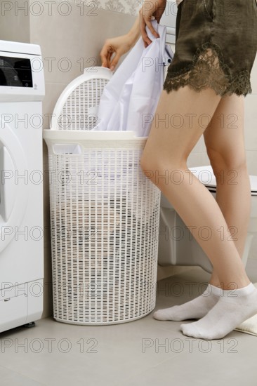 Unrecognizable woman stands next to a washing machine. She is putting white clothes into a laundry