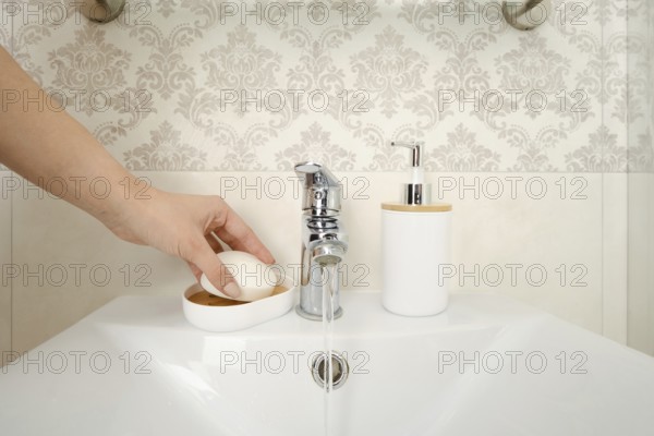 A hand reaches for a bar of soap placed on a dish near a sink. Water flows from a silver faucet