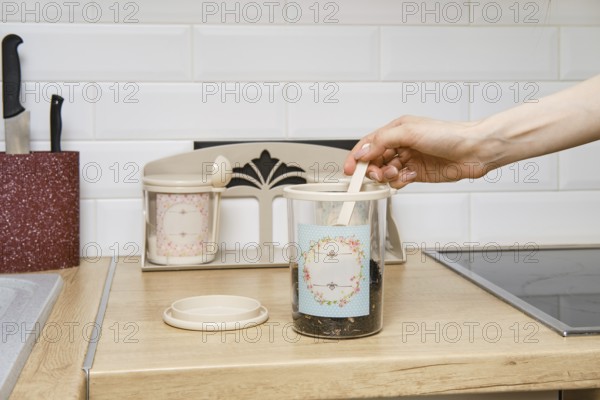 A hand is reaching into a jar with tea on a wooden kitchen countertop. Nearby, there is another jar