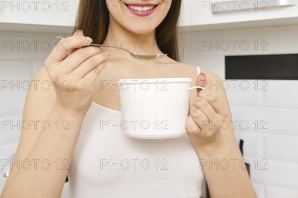 A woman stands in a kitchen holding large cup and spoon. She is smiling and appears to be preparing