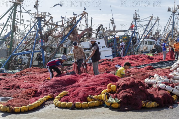 Men work on big red fishing nets, fishing boats in the background and seagulls in the sky