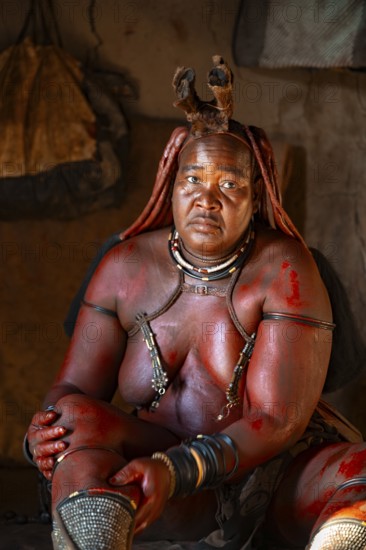 Elderly Himba woman rubbing herself with red paste for skin care, in a hut, traditional Himba